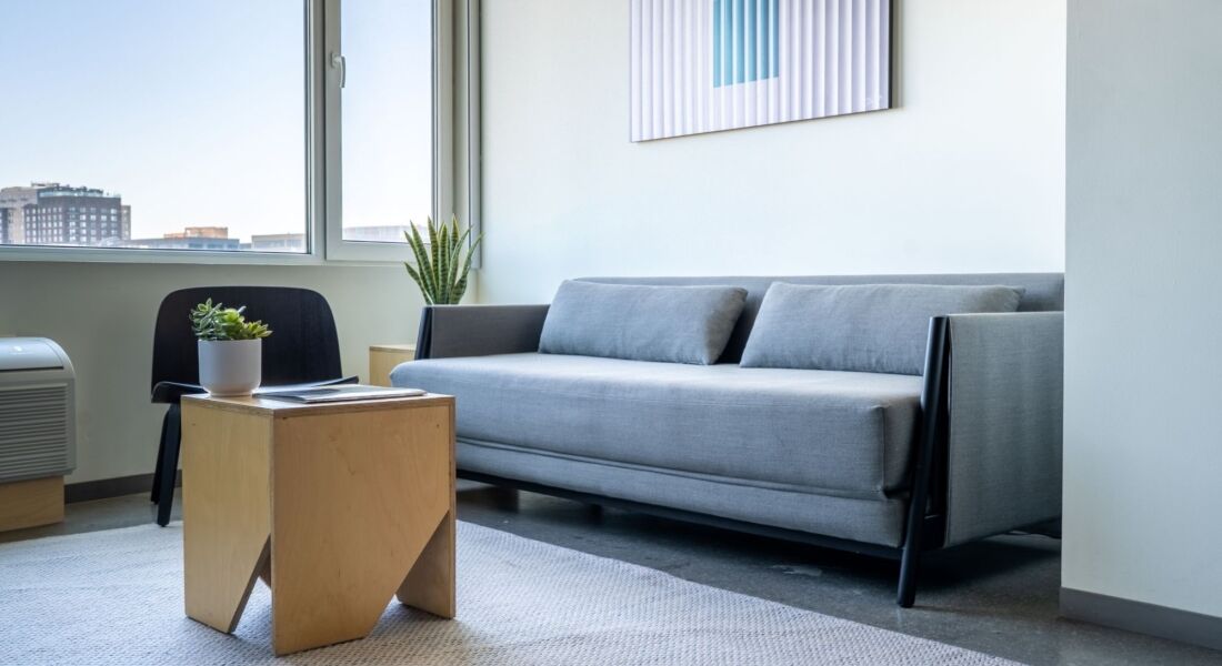 A modern lobby lounge with a gray sofa, wooden coffee table, black chair, and plants.