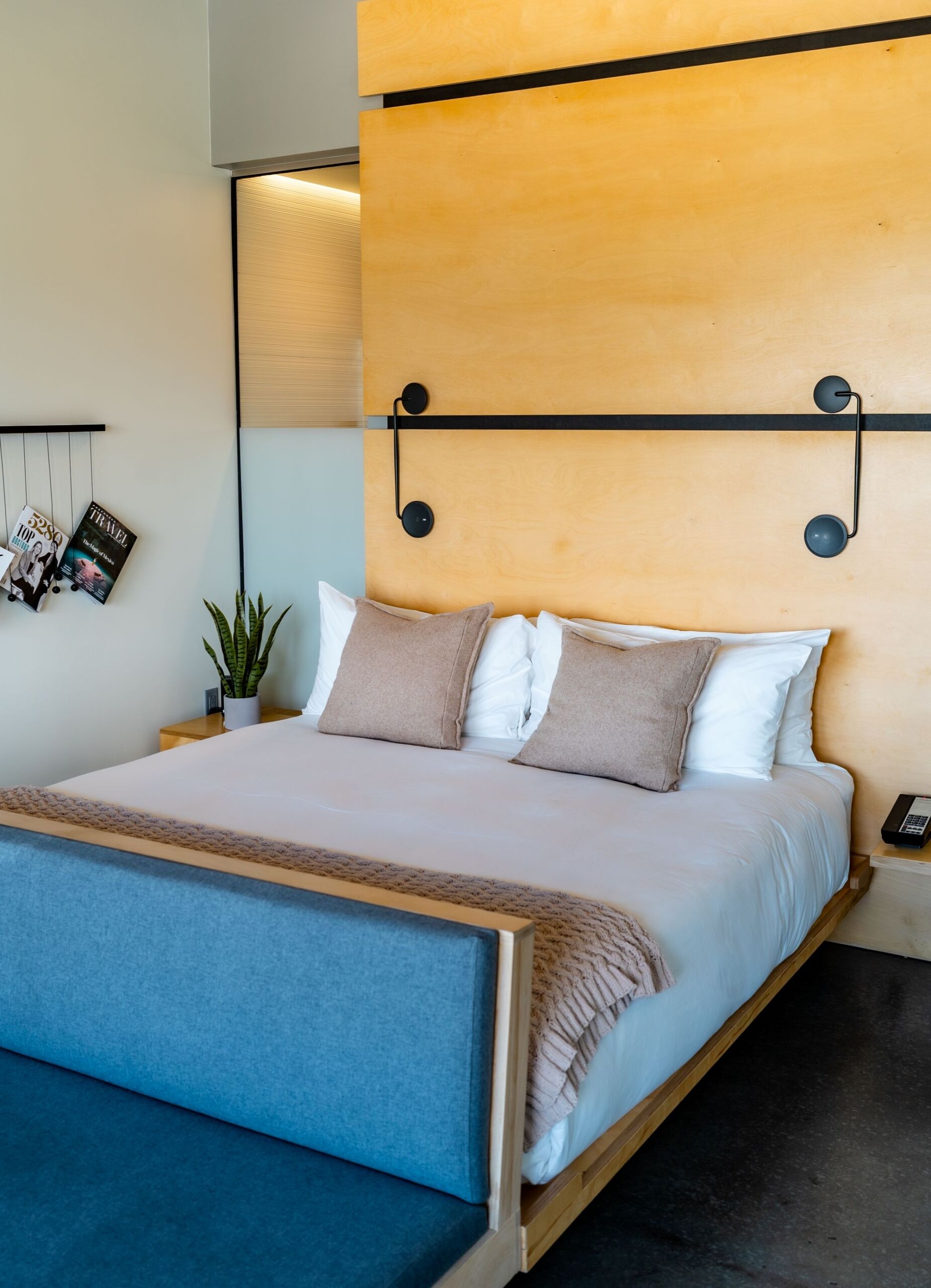 A neatly made king-sized bed with a blue upholstered footboard and a wooden headboard with black lights, in a room with wood paneling and reading materials.