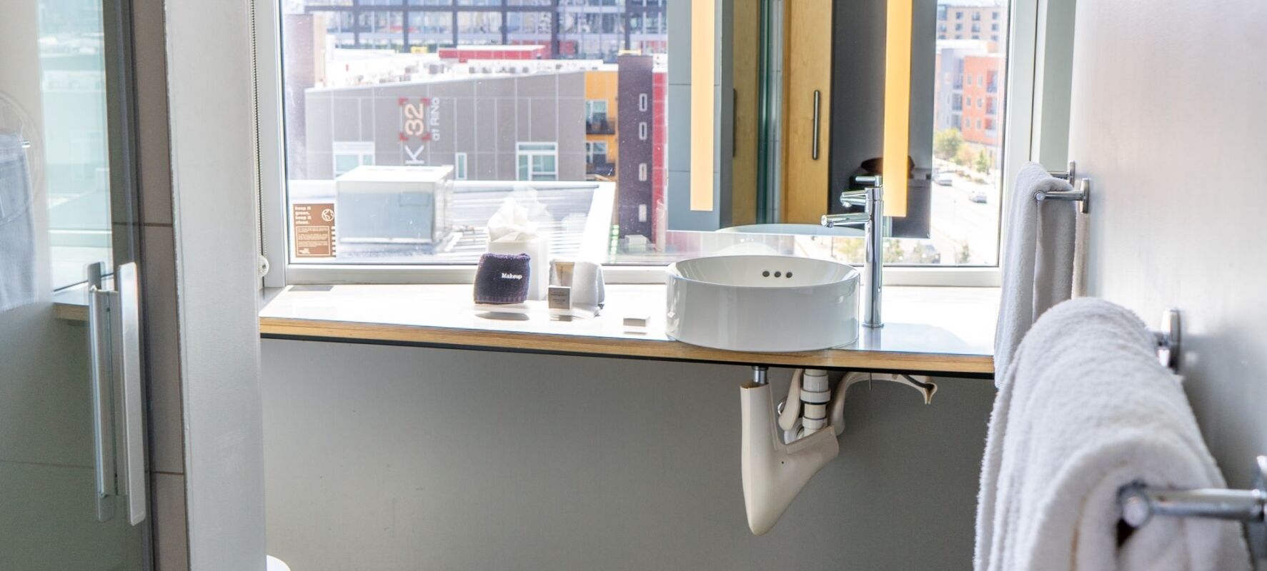 A bathroom sink area with a white sink, towels, and a window showing a city view.