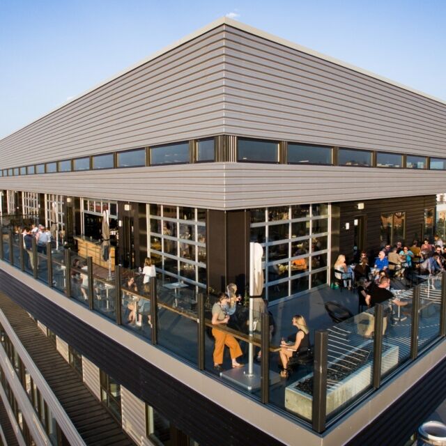 A rooftop bar with people sitting at tables, surrounded by glass railing and a modern building exterior.