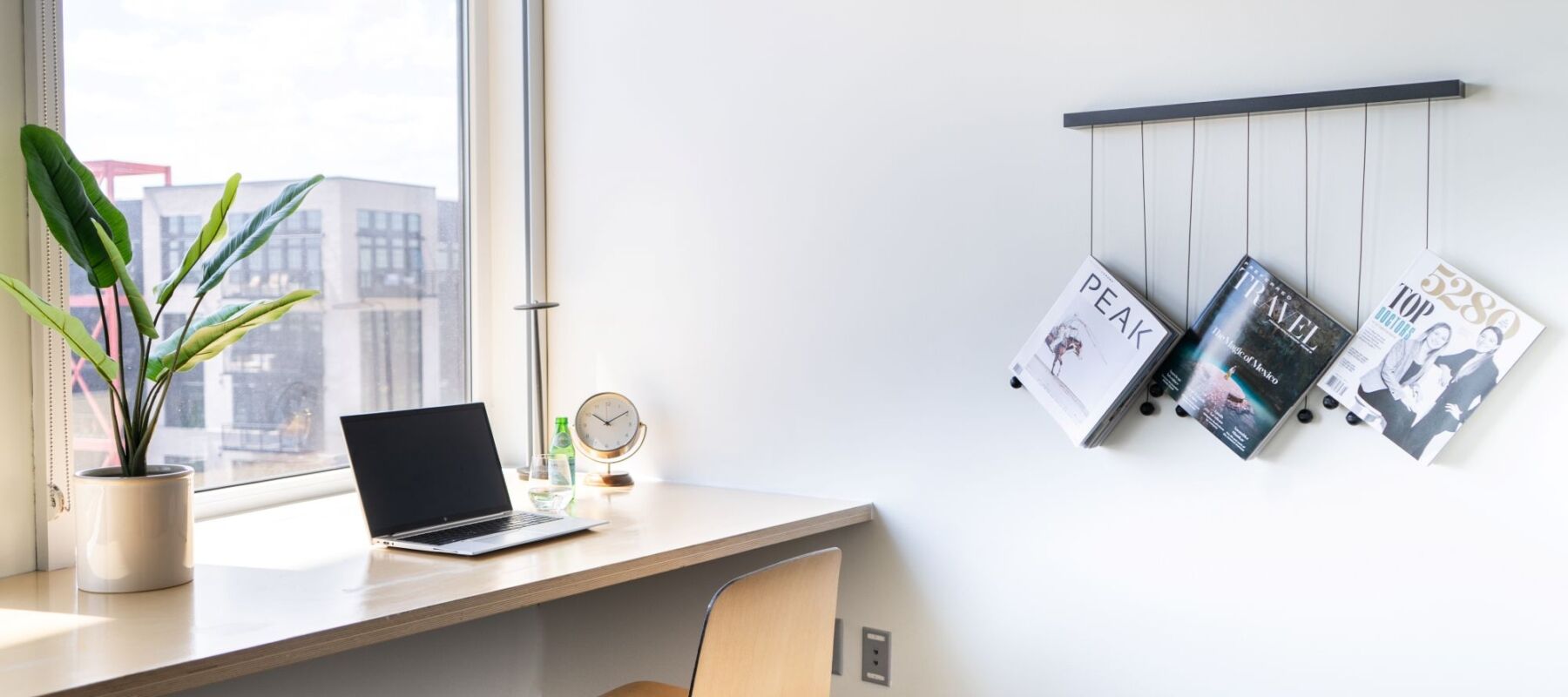 A well-lit workspace with a laptop, plant, and magazines near a window showing a city view.
