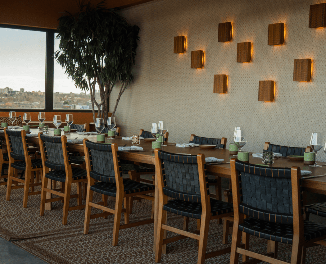 A long dining table with multiple chairs set for a meal, with a plant and wall lights in the background.