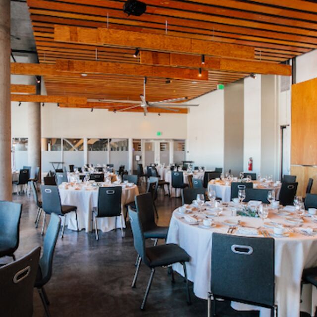 A large dining room with round tables set for a meal, white tablecloths, black chairs, and a wooden ceiling.