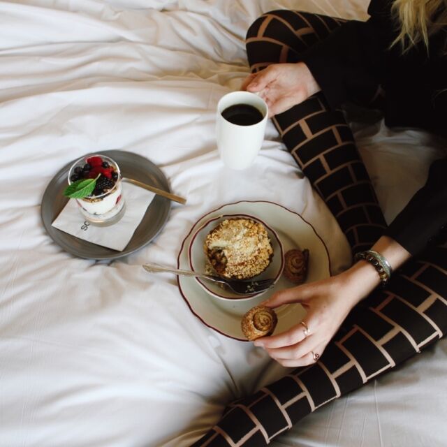 A woman is sitting on a bed with a breakfast tray and a cup of coffee.